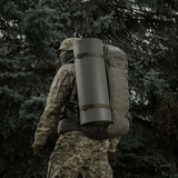 Hahmo camo-vaatteissa kantaa reppua, jossa M-Tac Liggeunderlag 20 mm (60x180 cm), metsäympäristössä.