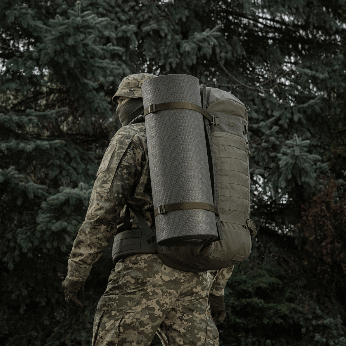 Hahmo camo-vaatteissa kantaa reppua, jossa M-Tac Liggeunderlag 20 mm (60x180 cm), metsäympäristössä.
