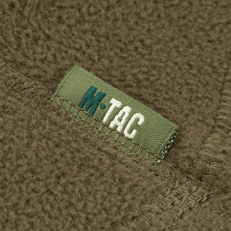 Lähikuva M-Tac Anatomisk Snood Justerbar Elite Fleece (320 g/m²) näkyvillä kangas-yksityiskohdilla ja logolla vihreällä taustalla.