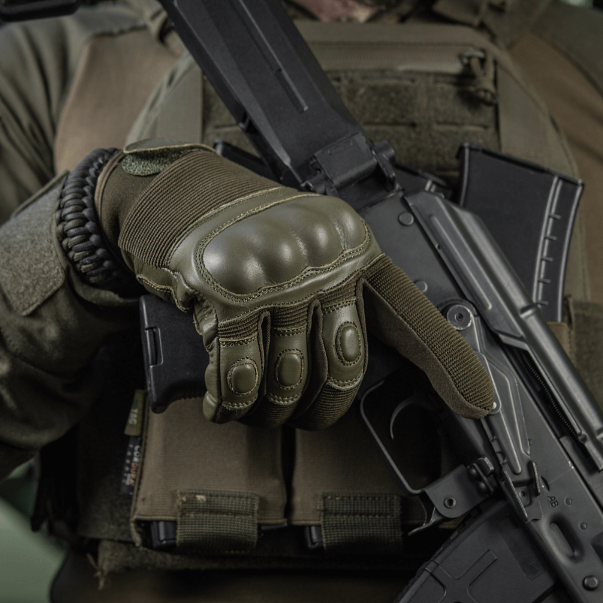 Henkilö pitelee asetta M-Tac Assault Tactical Mk.4 -hanskoissa; vahvistetut nyrkkipadat ja velcro turvalliseen istuvuuteen.