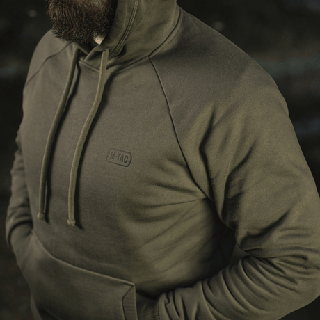 Mies pukeutuneena M-Tac Cotton Raglan Hard Hoodieen oliivinvärisenä, raglan-hihat, huppu ja kengurutasku. 100% puuvillaa fleece-vuorella lämmikkeeksi.