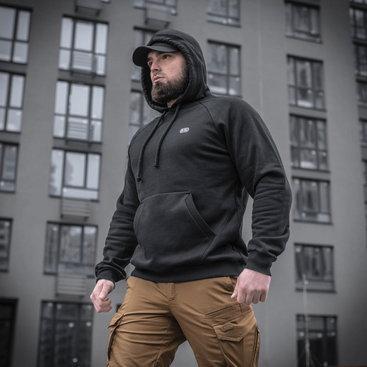 Mies mustassa M-Tac Cotton Raglan Hard Hoodie kengurutaskulla, seisoo ulkona rakennuksen edessä, sopii arkeen.