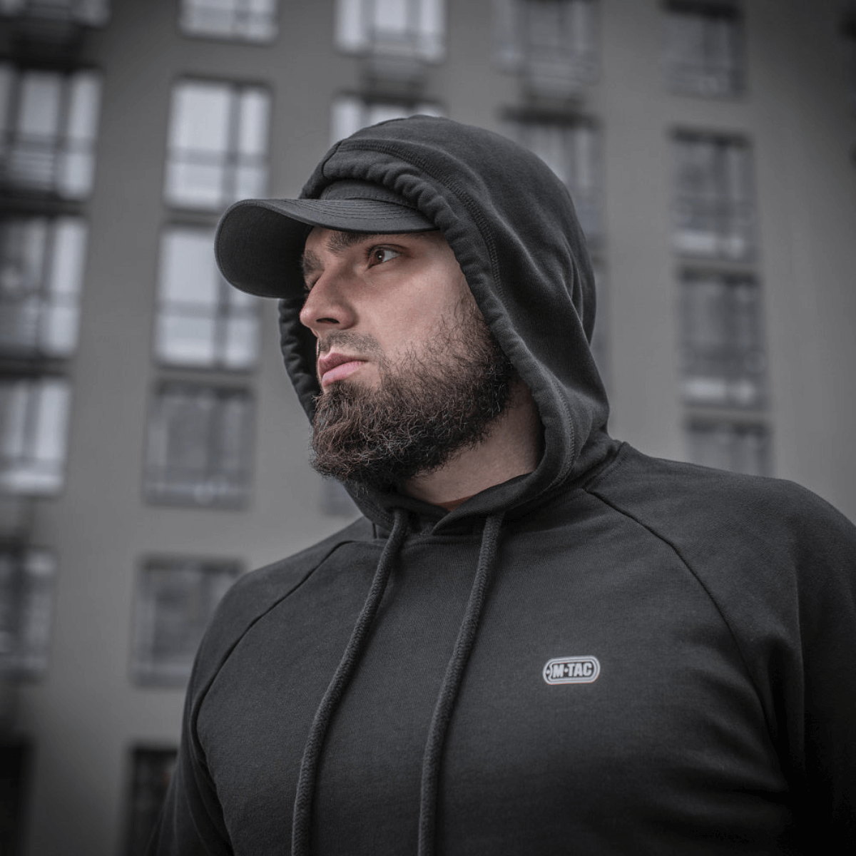 Mies pukeutuneena M-Tac Cotton Raglan Hard Hoodieen, 100% puuvillaa fleece-vuorella, tyylikäs arkipaita, nähty rakennuksen edessä.