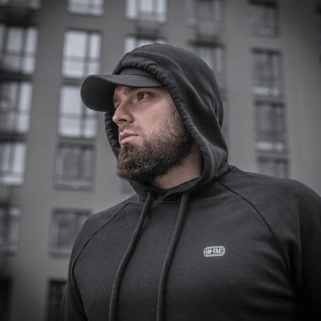 Mies pukeutuneena M-Tac Cotton Raglan Hard Hoodieen, 100% puuvillaa fleece-vuorella, tyylikäs arkipaita, nähty rakennuksen edessä.
