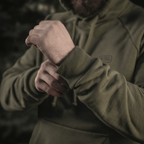 Henkilö pukeutuneena M-Tac Cotton Raglan Hard Hoodieen 100% puuvillaa fleece-vuorella vihreänä, näyttää joustavan hihansuun, sopii viileämpiin vuodenaikoihin.