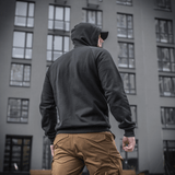 Mies M-Tac Cotton Raglan Hard Hoodiessa, 100% puuvillaa fleece-vuorella, sopii syksyyn ja talveen, nähty takaa rakennuksen edessä.