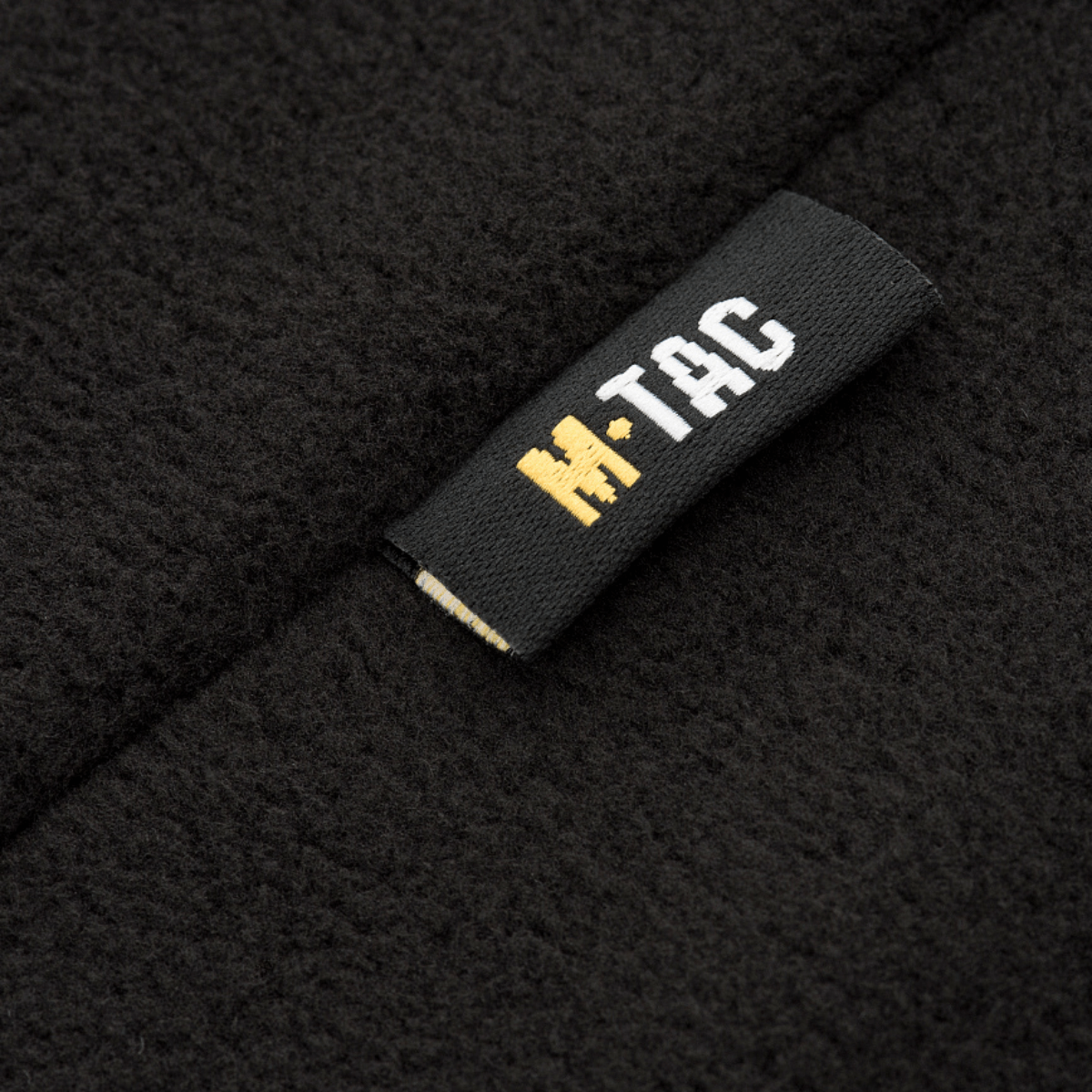 Lähikuva M-Tac Delta Fleece -takin etiketeistä, musta ja keltainen logo, fleece-materiaali, ihanteellinen ulkoiluun ja kylmiin olosuhteisiin