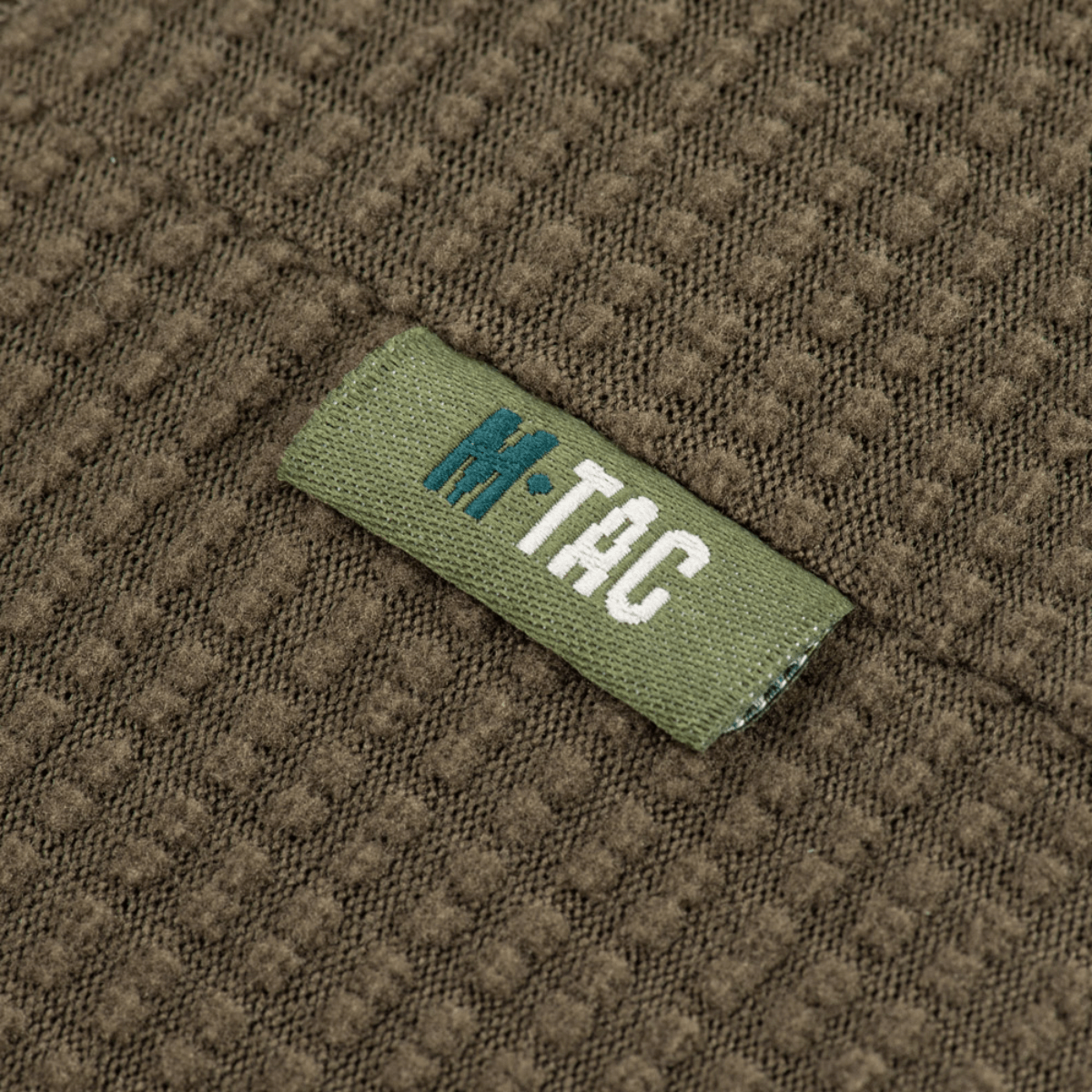Lähikuva M-Tac Hjelm Watch Cap Polartec -tekstiilirakenteesta M-Tac-logolla, ihanteellinen kylmään säähän ja mukavuutta kypärän alla.