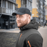 Miehellä on M-Tac Mesh Flex Ripstop Baseball Cap modernien rakennusten edessä, täydellinen ulkoiluun, musta lippis mesh-paneeleilla.