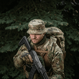 Mies maastoverkossa M-Tac Panama Hat Gen.II NYCO päässään, taktinen univormu ja reppu metsäympäristössä.