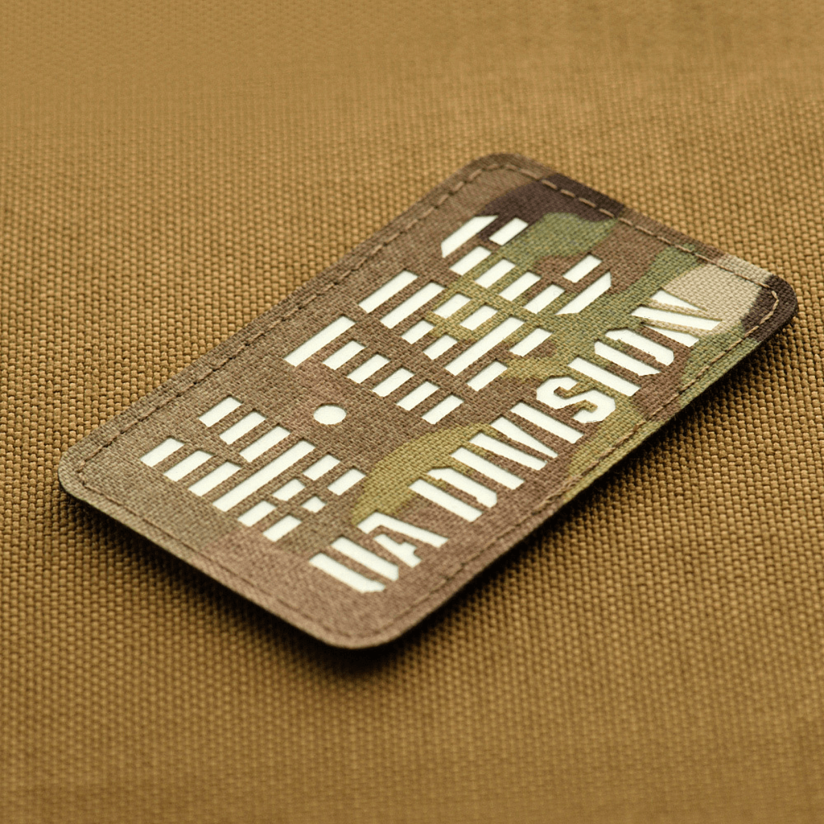 M-Tac Patch UA Division Laser Cut pimeässä hohtavalla materiaalilla ja Velcro-kiinnityksellä, Cordura 500D-materiaalia, ruskealla taustalla.