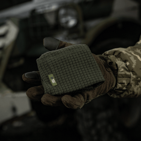M-Tac Rip-Stop Fleece Hjelmliner esitetty kädessä, jossa taktinen hanska, ulkoilmamaisessa sotilasympäristössä, kompakti ja käyttövalmis.