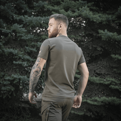 Mies M-Tac Sniper T-paidassa metsäympäristössä takaa, näyttää istuvuuden ja tiukan leikkauksen