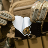 Henkilö taktisia hanskoja käyttäen pitää M-Tac Sticker Valkyrie -tarran laukun edessä.