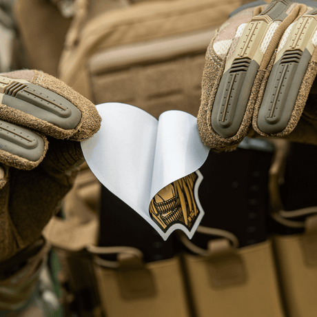 Henkilö taktisia hanskoja käyttäen pitää M-Tac Sticker Valkyrie -tarran laukun edessä.