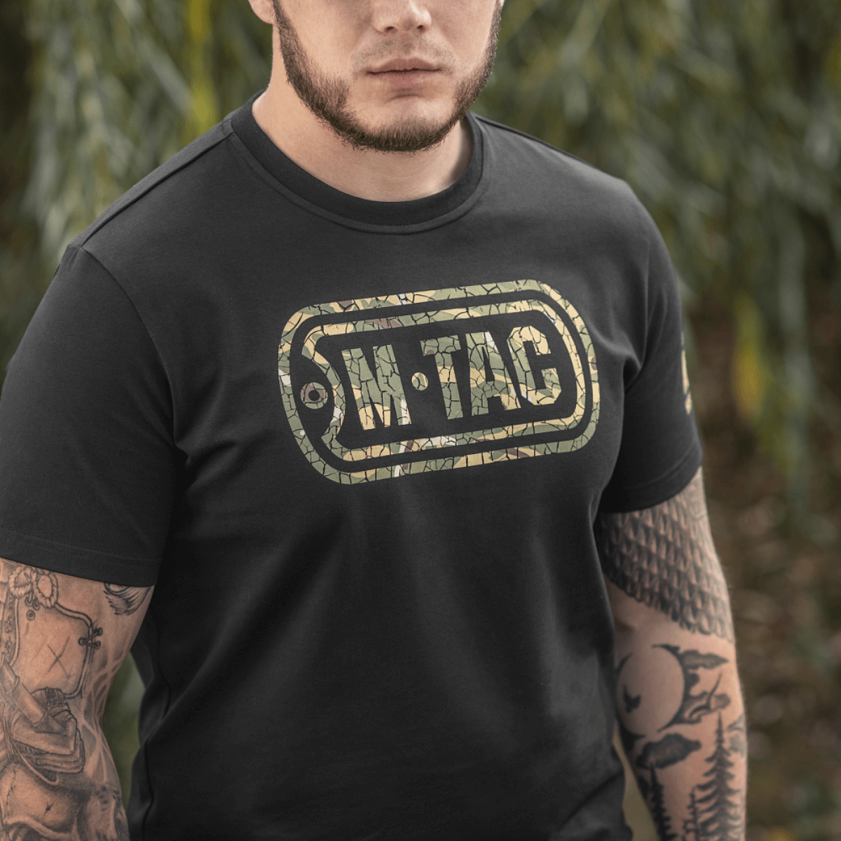 Mies M-Tac T-Shirt Logossa, camo, hihan slogan ja "Military&Tactical Since 2014" selässä, ympäröity vihreillä kasveilla.