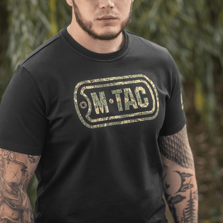 Mies M-Tac T-Shirt Logossa, camo, hihan slogan ja "Military&Tactical Since 2014" selässä, ympäröity vihreillä kasveilla.