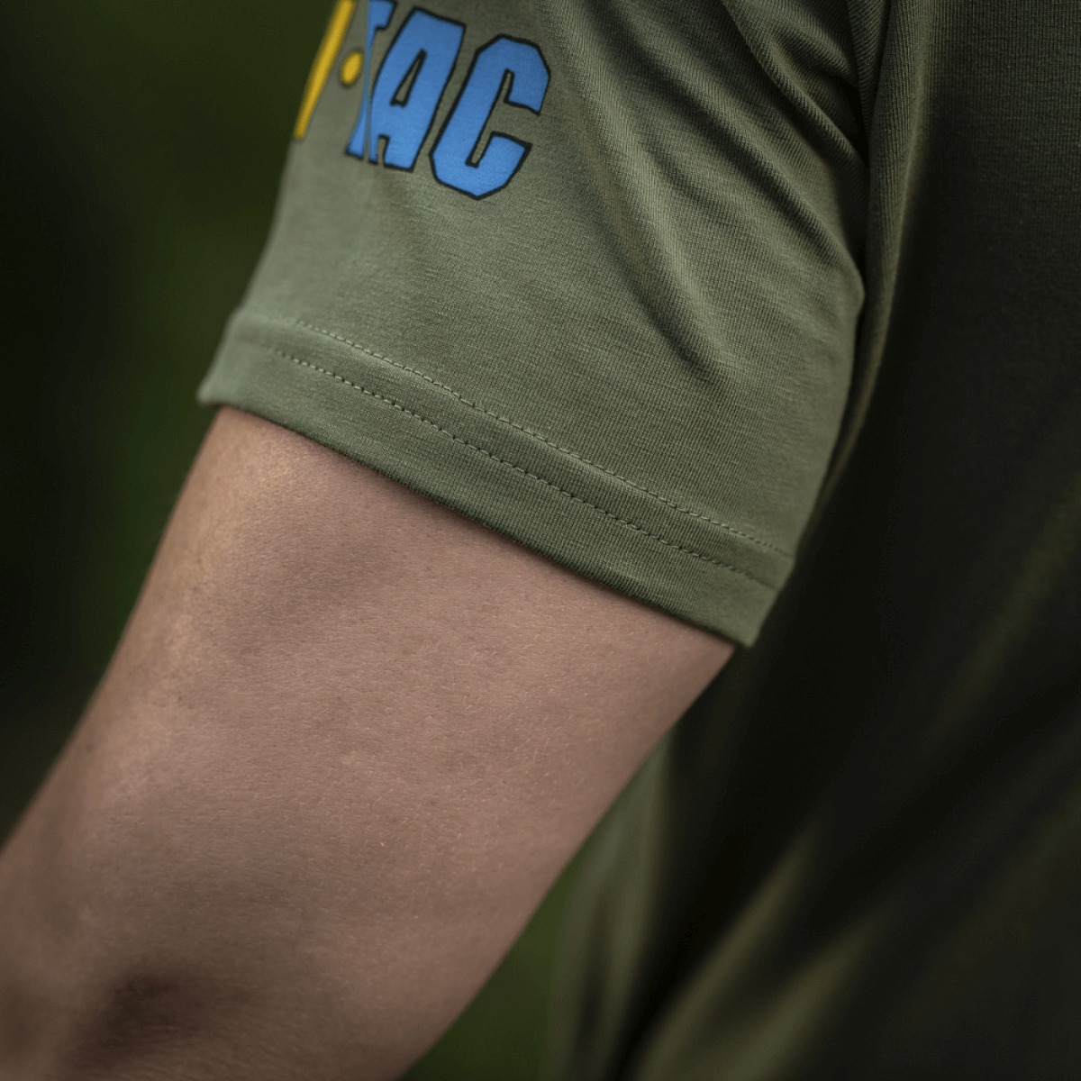 Lähikuva hihasta M-Tac T-Shirt Quadcopter, logo sinikeltaisena oliivinvihreällä taustalla, näyttää puuvilla-elastaani-seoksen.