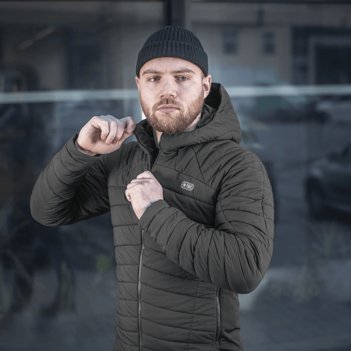 Mies pukeutuneena M-Tac Jarl -takkiin rakennuksen edessä, säätää takkia ja katsoo mietteliäänä eteenpäin urbaanissa ympäristössä.
